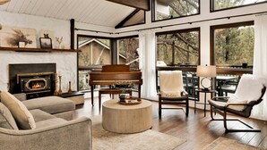 A spacious living room with a fireplace, a grand piano, and large windows offering views of the outdoors. - Soaring Pines #2193 (Big Bear Lake)