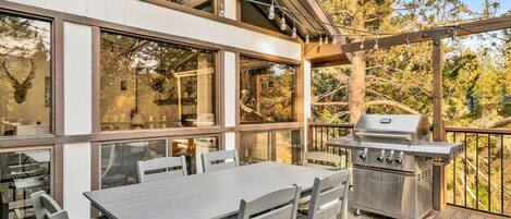 An outdoor patio with a table and chairs, a grill, and large windows overlooking trees.