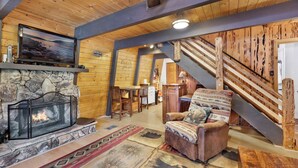 Un salon chaleureux avec une cheminée en pierre, des murs en bois, un fauteuil confortable et un escalier menant à un autre niveau.