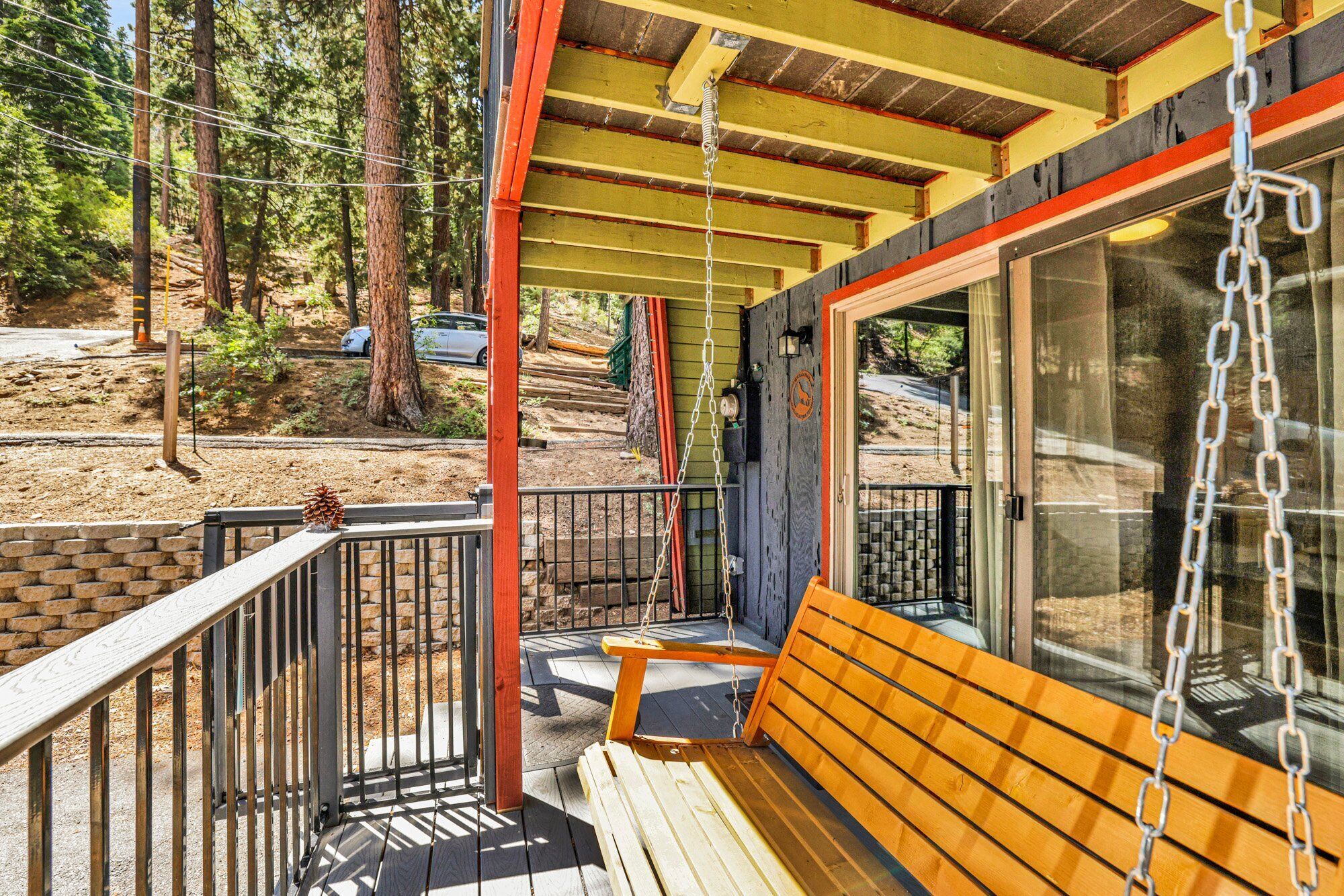 Une terrasse en bois avec une balançoire, un espace couvert doté d’une rambarde en métal et une vue sur une zone boisée.