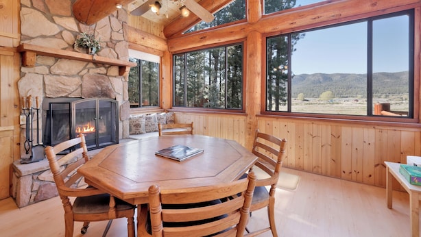 A wooden interior with a stone fireplace, a dining table with chairs, and large windows offering a view of the outdoors. - THE Alps Lodge #2142 (Big Bear Lake)