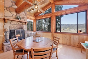 Un interior de madera con chimenea de piedra, una mesa de comedor con sillas y amplias ventanas que ofrecen vistas al exterior.