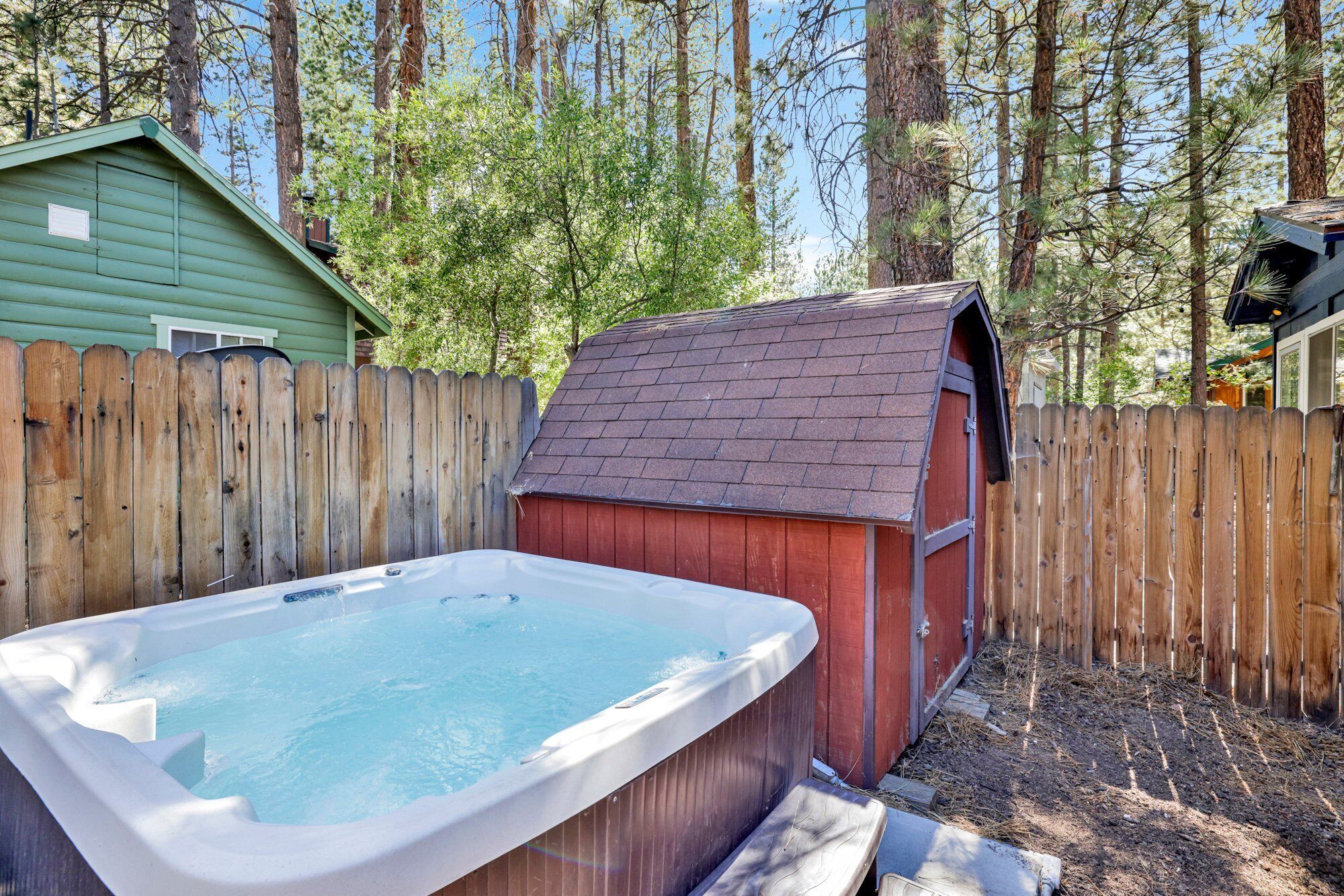 Un jacuzzi en un patio trasero con una cerca de madera, un cobertizo y árboles al fondo.