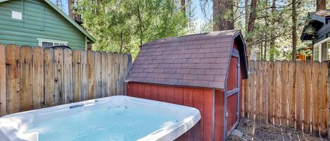 Un jacuzzi en un patio trasero con una cerca de madera, un cobertizo y árboles al fondo.