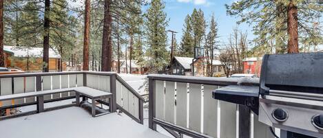 A snow-covered deck with a grill and a basketball hoop, surrounded by tall pine trees and houses in a residential area.