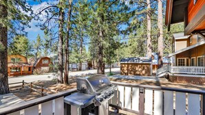 A deck with a grill, surrounded by tall pine trees and a cabin-style building. - Bearadise Cottage #2275 (Big Bear Lake)