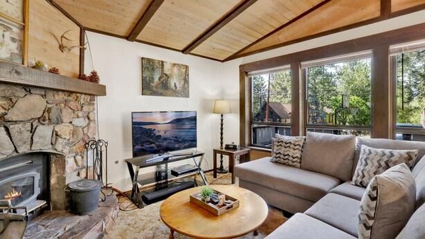 A cozy living room with a stone fireplace, a large TV, comfortable seating, and a view of the outdoors through large windows. - Bearadise Cottage #2275 (Big Bear Lake)