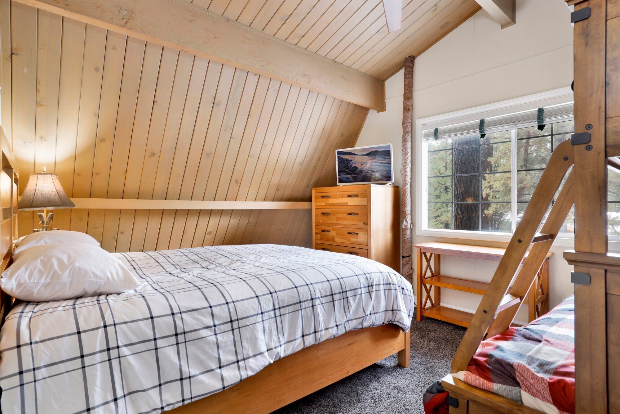 Une chambre avec un plafond en pente, un lit recouvert d’une couverture à carreaux, une commode en bois, un bureau et une fenêtre donnant sur des arbres.