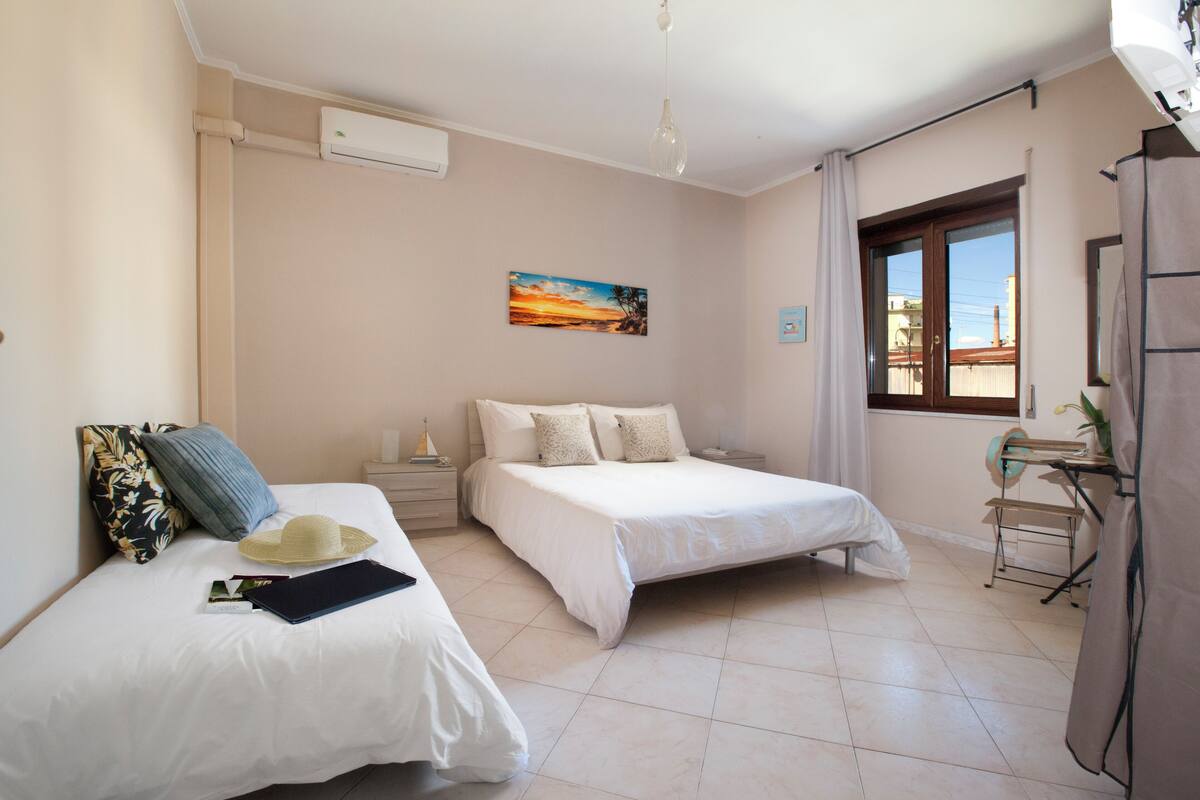 Una camera da letto con due letti, un comodino, una finestra con tende, un quadro al muro e una ventola a soffitto.