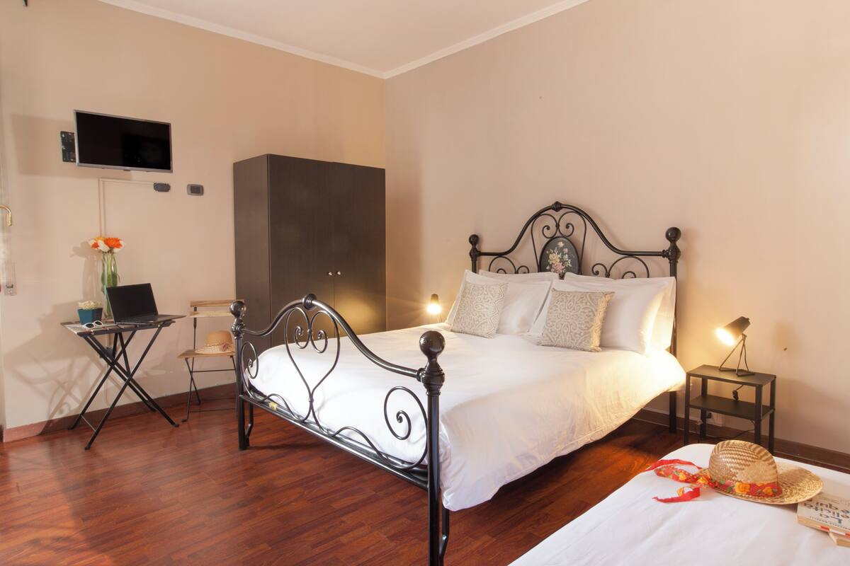 Una camera da letto ordinata con una struttura letto in metallo, un comodino con una lampada, un portatile su un tavolino e un cappello sul letto.
