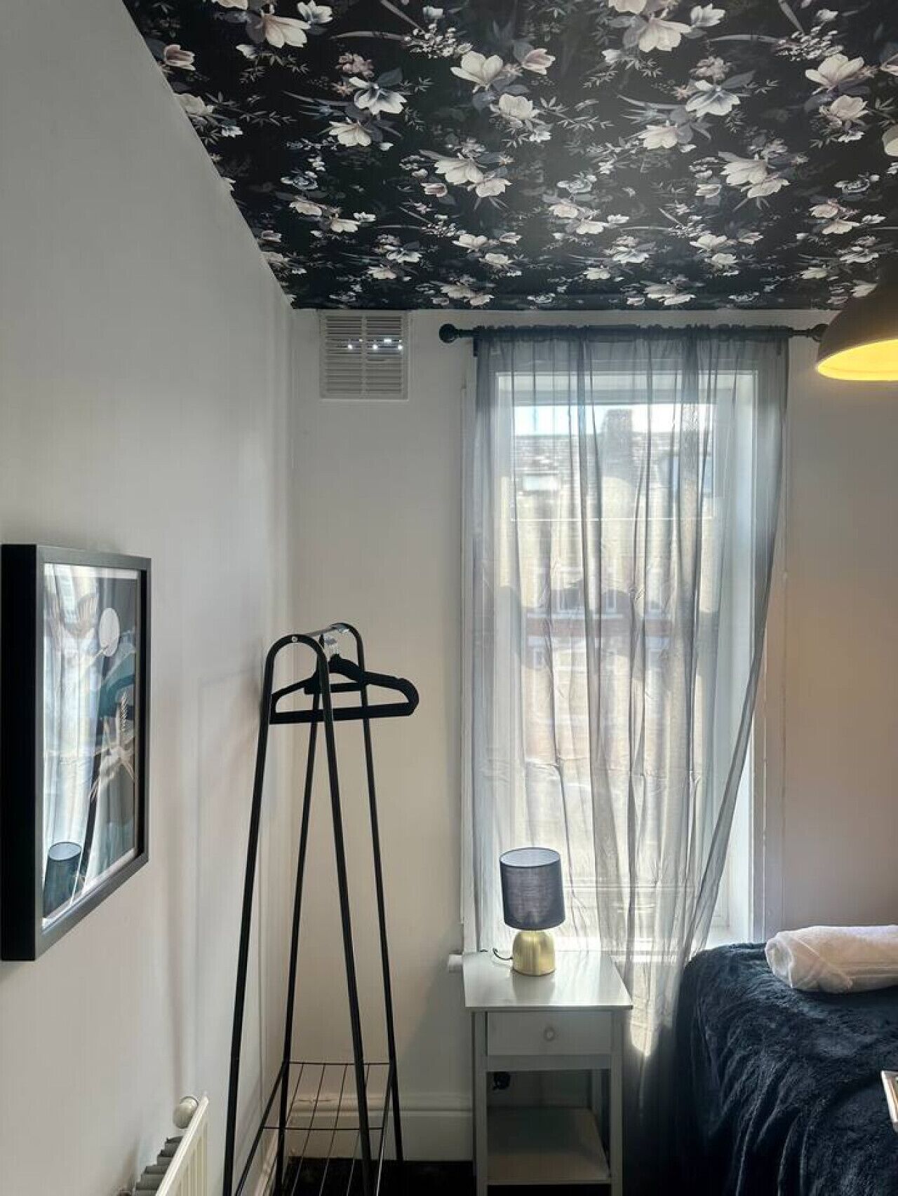 Un dormitorio con cama, una mesita de noche con lámpara, perchero, ventana con cortinas translúcidas y pared con estampado floral.