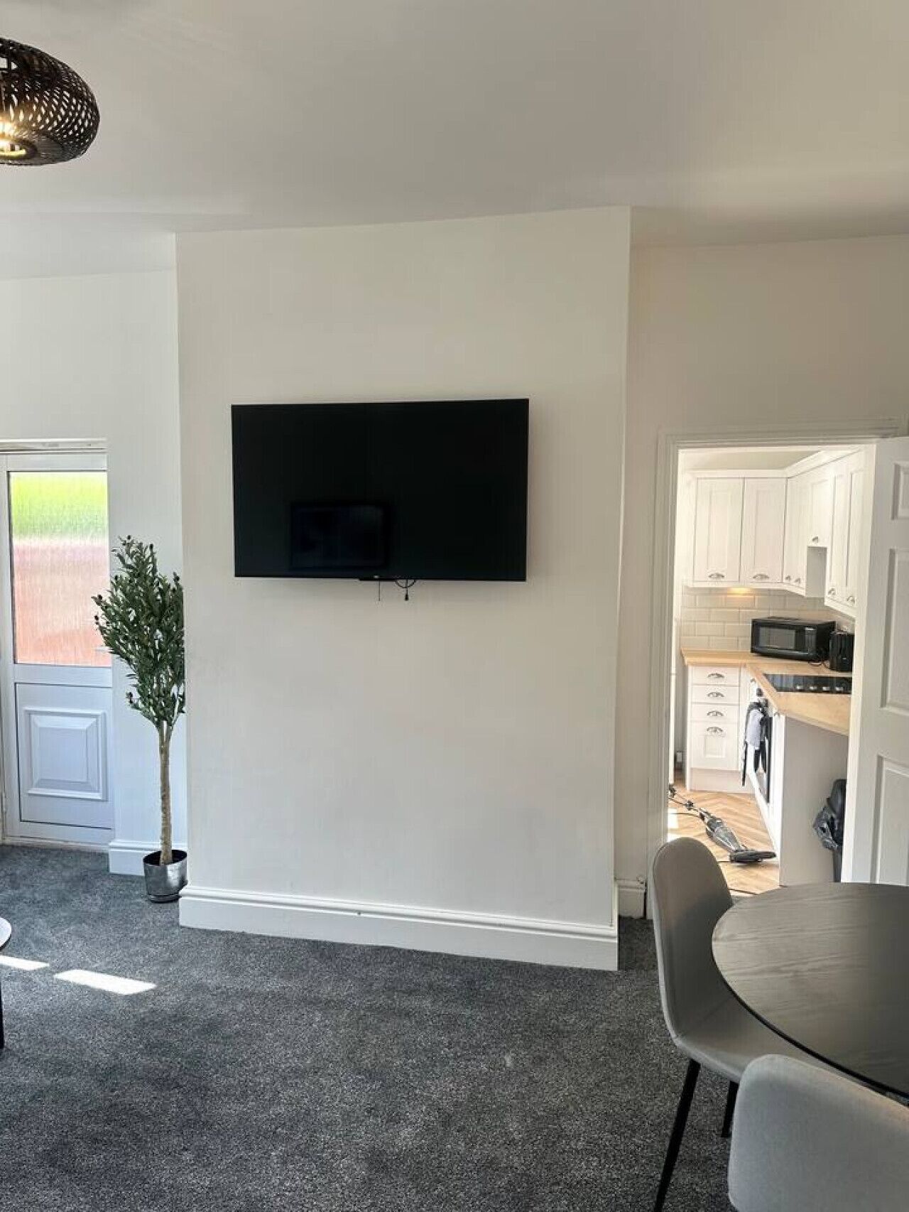 A modern living room with a flat-screen TV mounted on the wall, a dining area with a round table and chairs, and a kitchen visible through an open doorway.