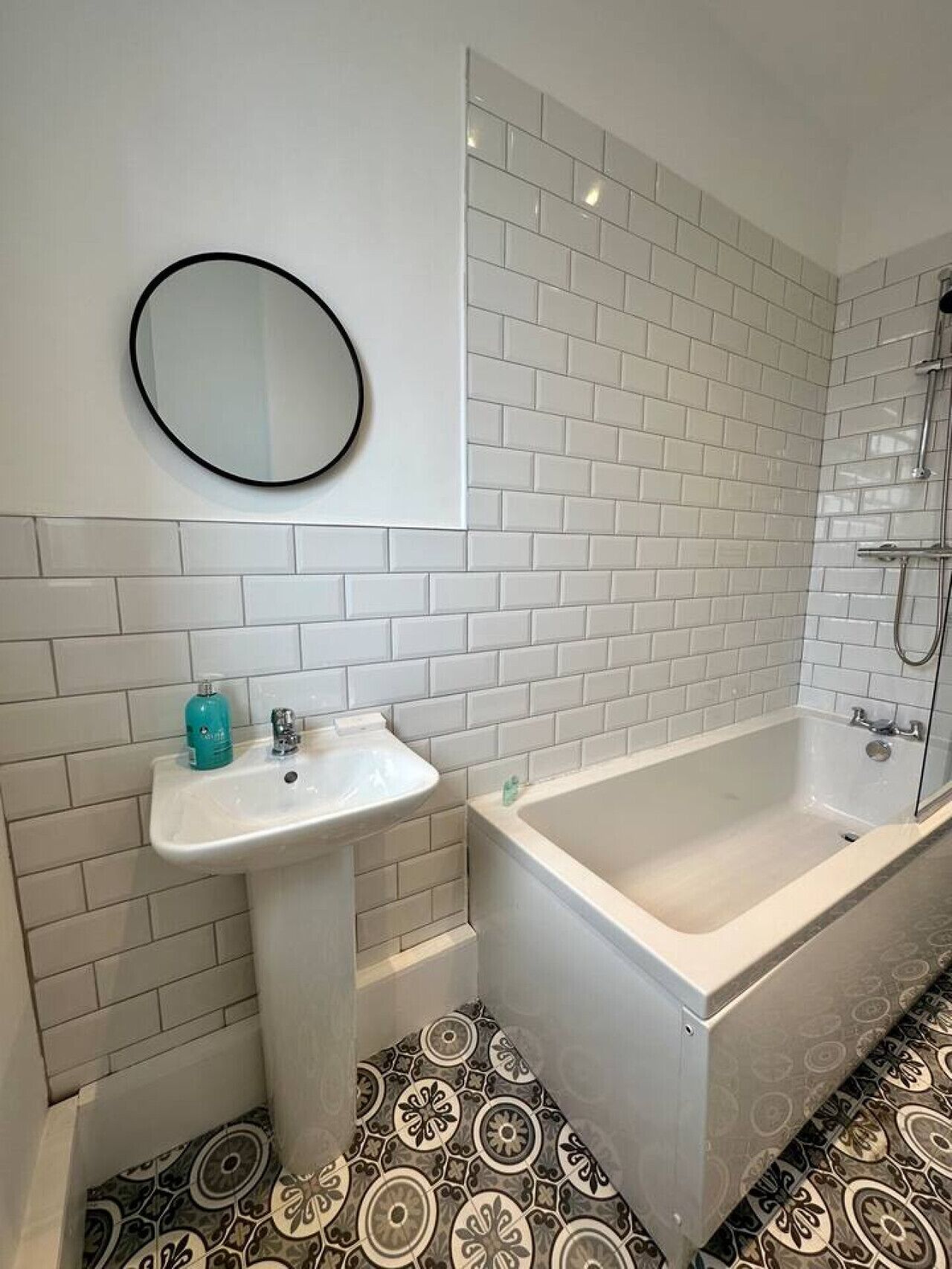 A bathroom with white subway tiles, a pedestal sink, a bathtub, a shower, and a patterned floor.