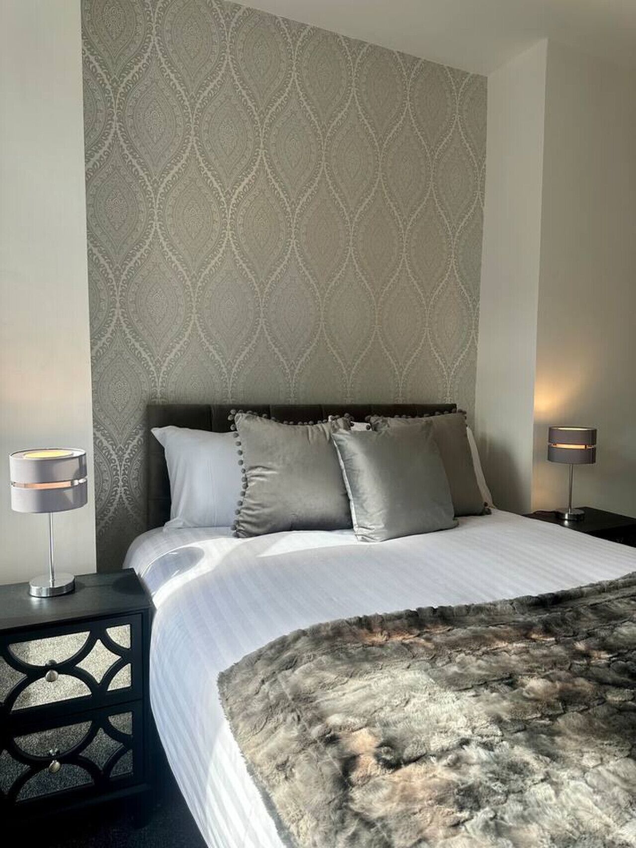 A neatly made bed with a grey blanket, two bedside lamps, and a patterned wallpaper.