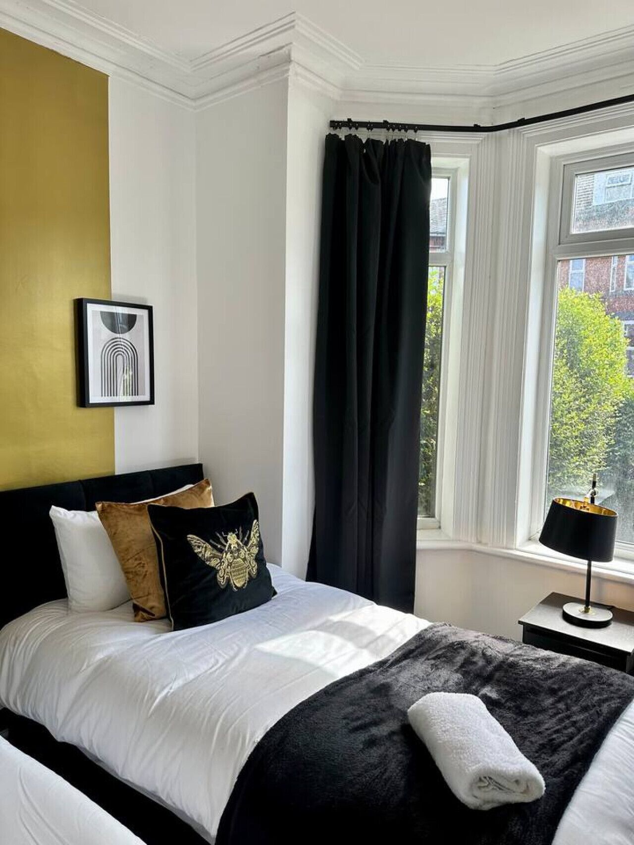 Een hotelkamer met een bed, een raam met gordijnen, een ingelijst schilderij aan de muur en een nachtkastje met een lamp.