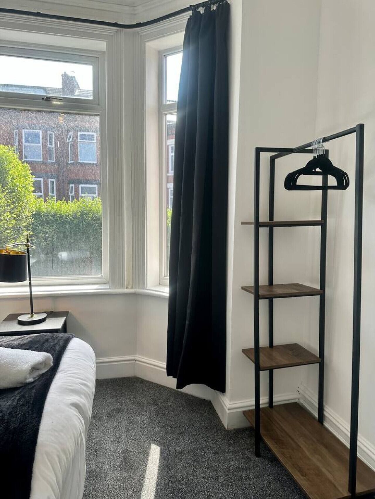 Un dormitorio con una cama, un estante negro para colgar ropa, una ventana con vista a edificios y una planta en maceta.