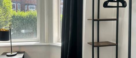 Un dormitorio con una cama, un estante negro para colgar ropa, una ventana con vista a edificios y una planta en maceta.