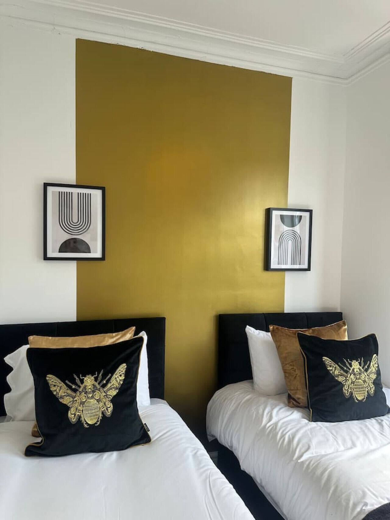 A hotel room with two beds, each with black and gold pillows, and a large gold accent wall.