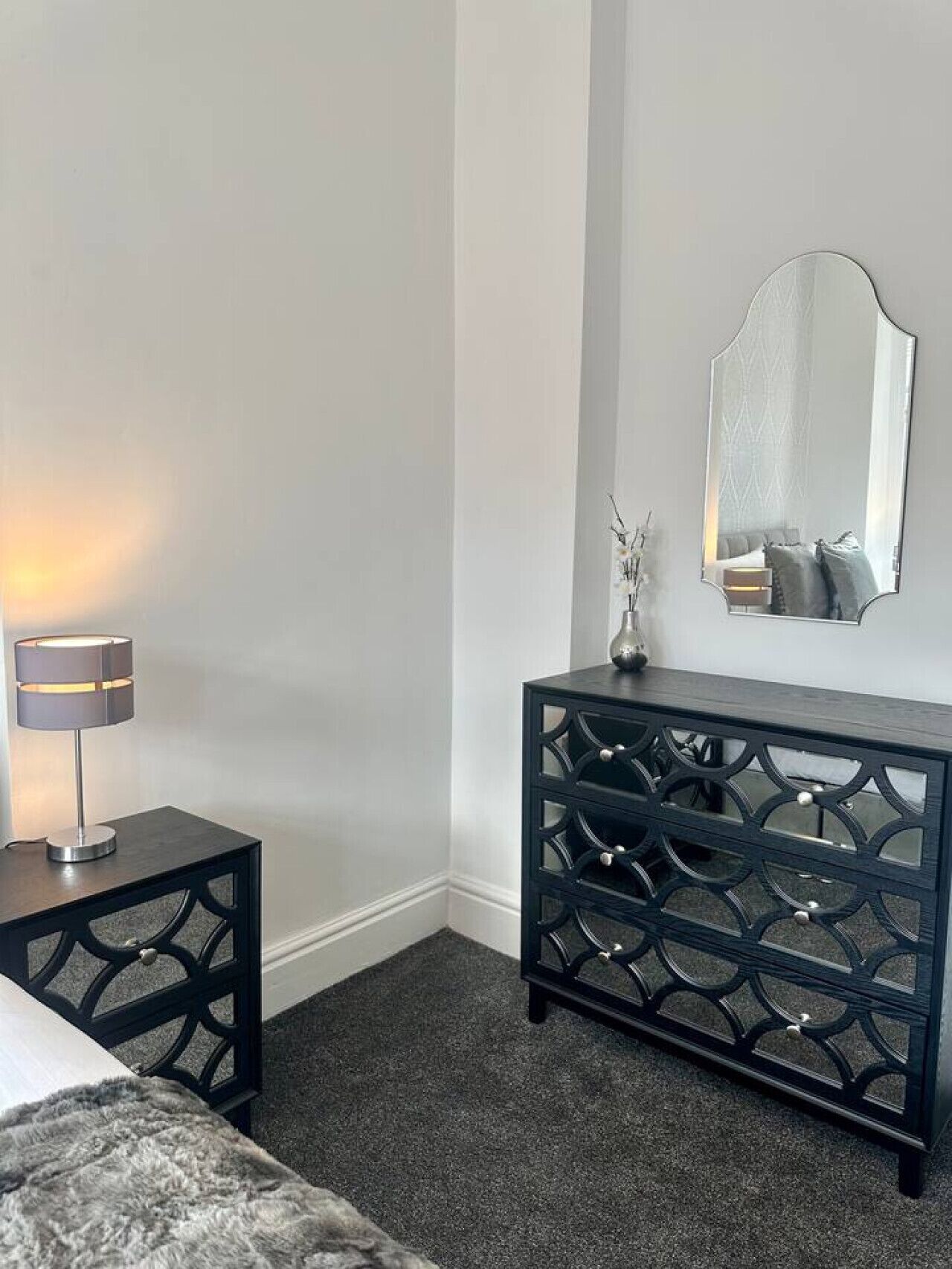 A bedroom with a bed, a bedside table with a lamp, a dresser, and a mirror on the wall.