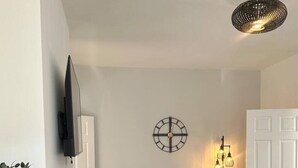 Ein Essbereich mit einem runden Tisch, vier Stühlen, einer Wanduhr, einer Stehlampe und einem an der Wand befestigten Fernseher.