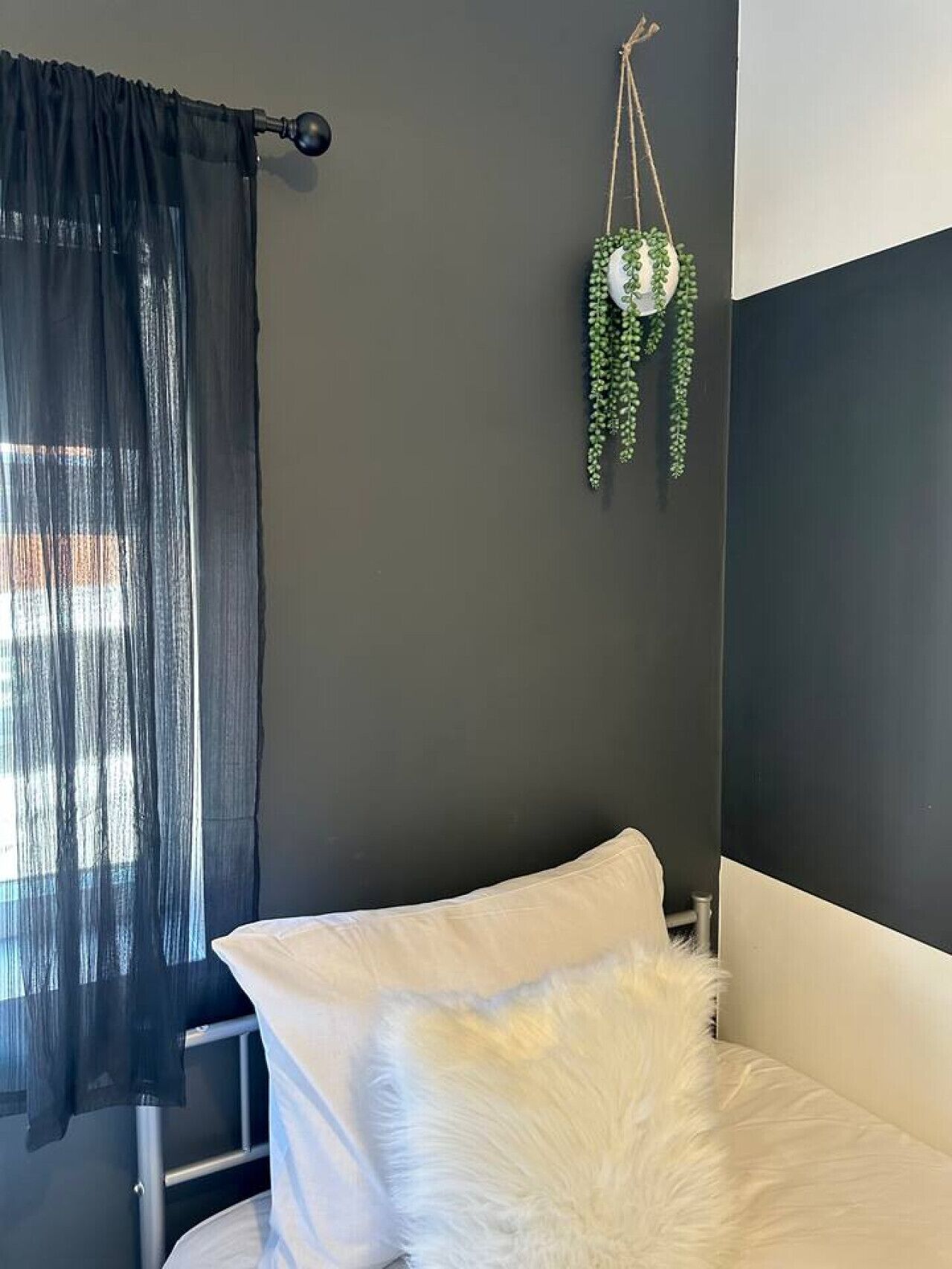 A bedroom with a bed, a hanging plant, and a window with sheer curtains.