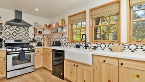 Una cocina moderna con armarios de madera, estufa de acero inoxidable y lavavajillas. Hay ventanas con persianas y vistas a árboles.