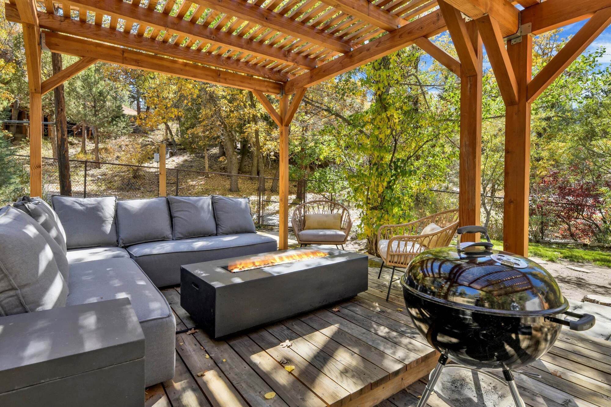 Une terrasse extérieure dotée d’une pergola en bois, d’un canapé d’angle gris, d’un barbecue rond et d’un fauteuil en osier.