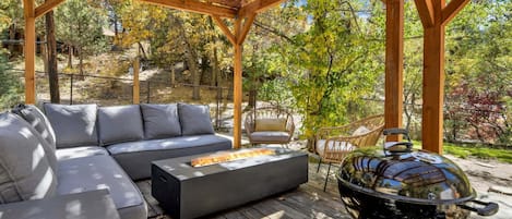 Un patio exterior con una pérgola de madera, un sofá seccional gris, una parrilla redonda y una silla de mimbre.