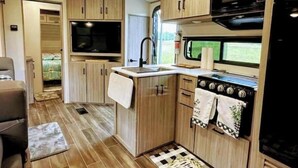 A compact RV kitchen with wooden cabinets, a microwave, a stove, and a sink. - City-Accessible Camper (Goldston)