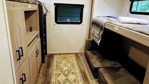 A compact RV interior with a bunk bed, a small window, and a storage cabinet. - City-Accessible Camper (Goldston)
