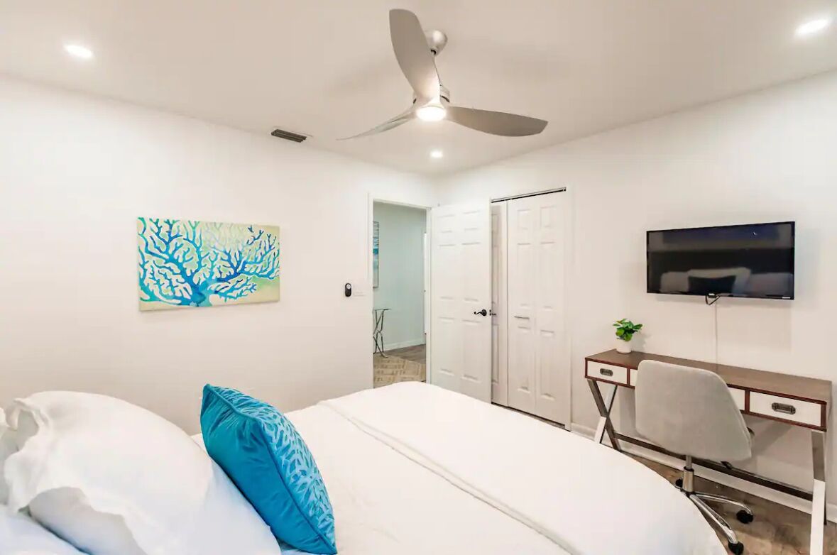 Une chambre à coucher avec un lit, un bureau et une chaise, un ventilateur de plafond, une télévision et une porte.