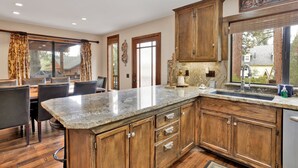 A modern kitchen with wooden cabinets, a granite countertop, and a dining area with leather chairs. - Luxury Lodge #1888 (Big Bear Lake)