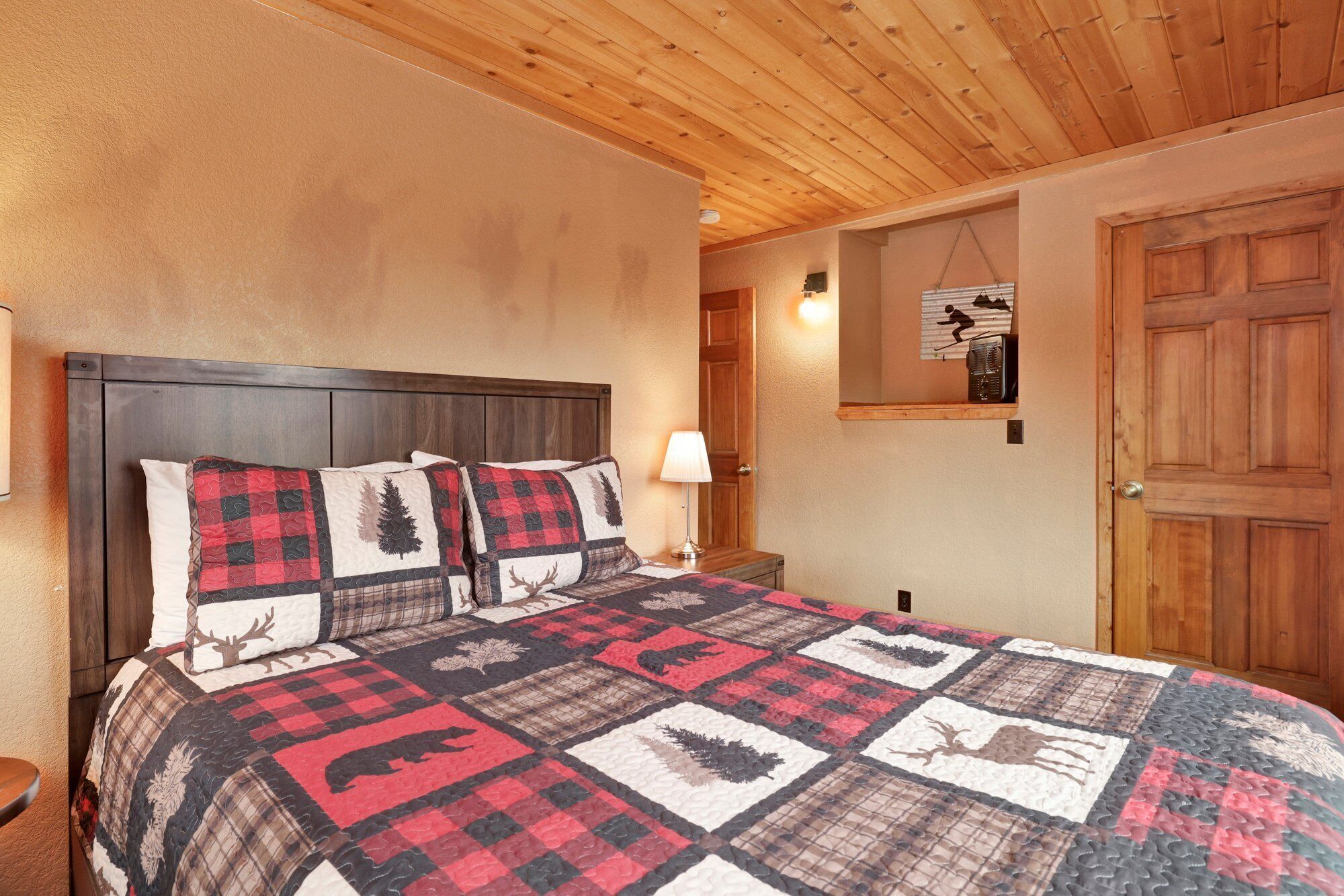 Quarto com teto de madeira, cama com colcha estampada com animais, mesa de cabeceira com abajur e porta com foto de uma pessoa esquiando.