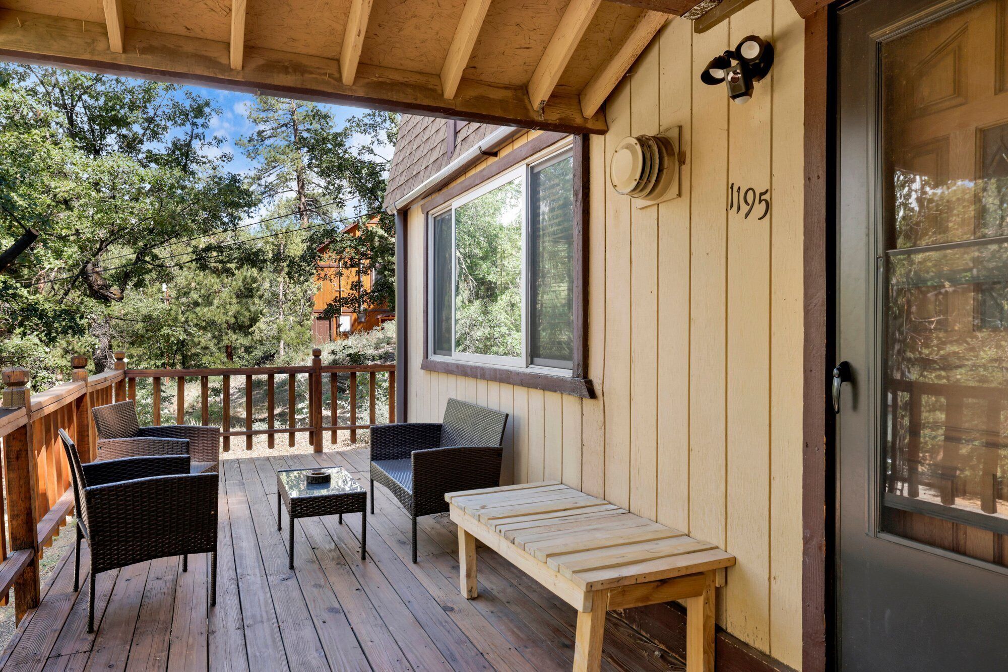 Una terraza de madera con mobiliario de mimbre, un banco y una puerta con el número 1195.