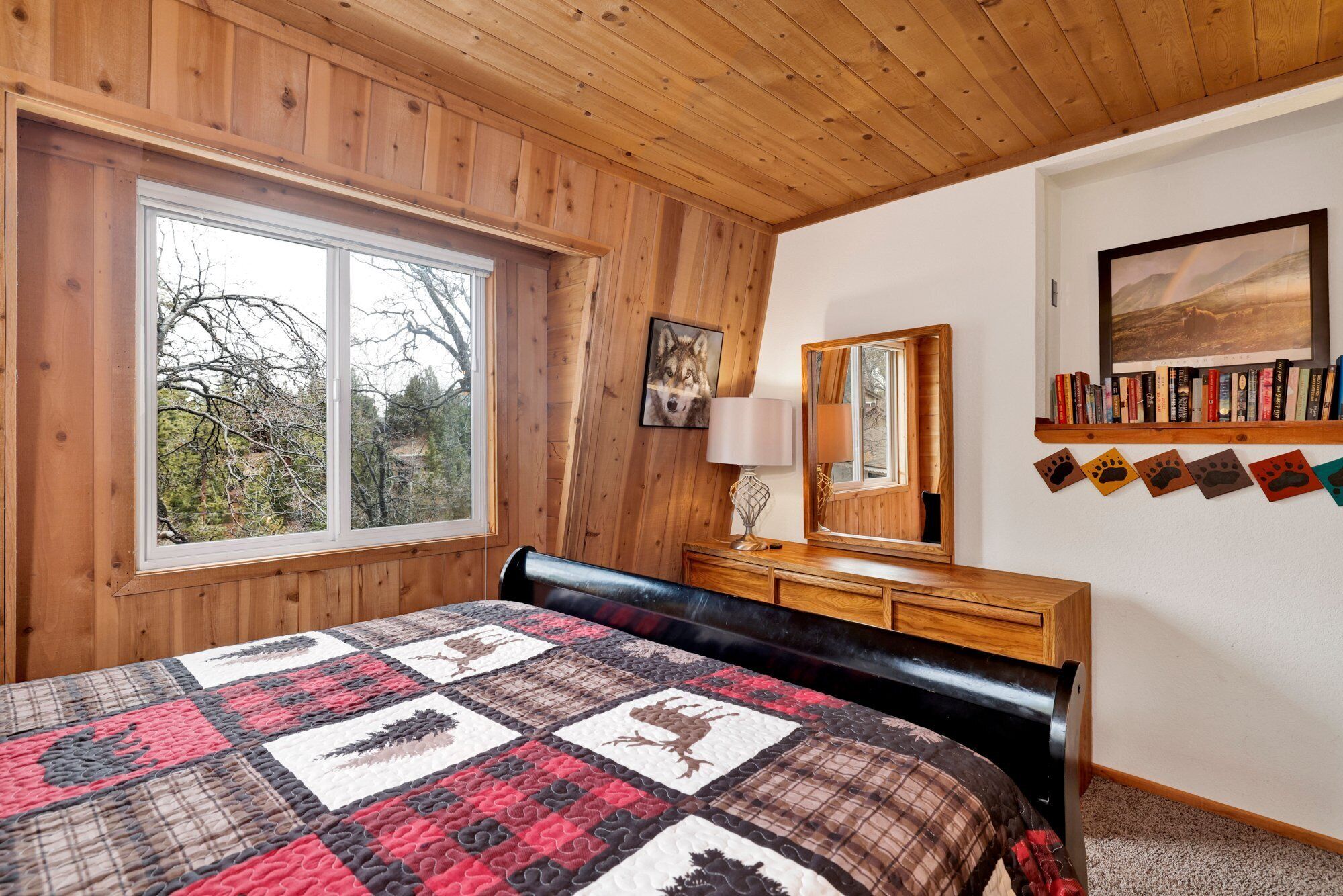 A bedroom with a large window, a wooden headboard, a quilted bedspread, a bedside table with a lamp, a mirror, and a bookshelf with books.