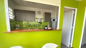 A kitchen with a green wall, a wooden countertop, and a clock. There are two white chairs and a microwave in the background. - VILLA ANAIS sea view, Trois Ilets, quiet, swimming pool, close to shops  (LES TROIS ILETS)
