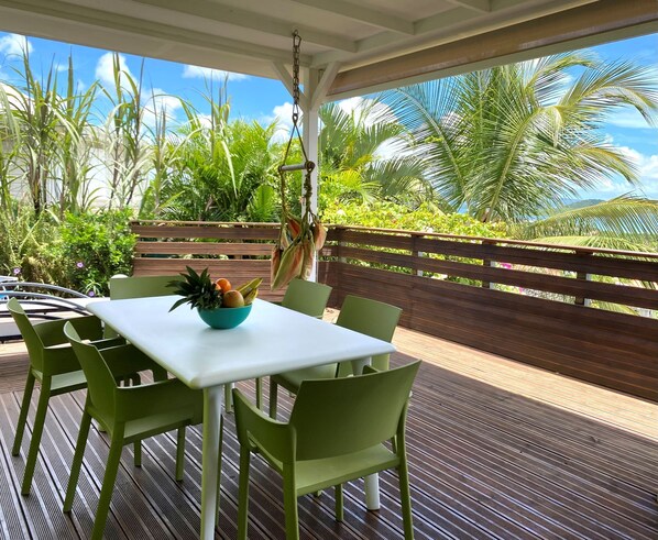 An outdoor seating area with a table and chairs, a bowl of fruit, and a view of tropical plants and a wooden fence.