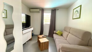 A living room with a beige sofa, a wooden coffee table, a TV on a white cabinet, a window with curtains, and a wall-mounted air conditioner. - VILLA ANAIS sea view, Trois Ilets, quiet, swimming pool, close to shops  (LES TROIS ILETS)