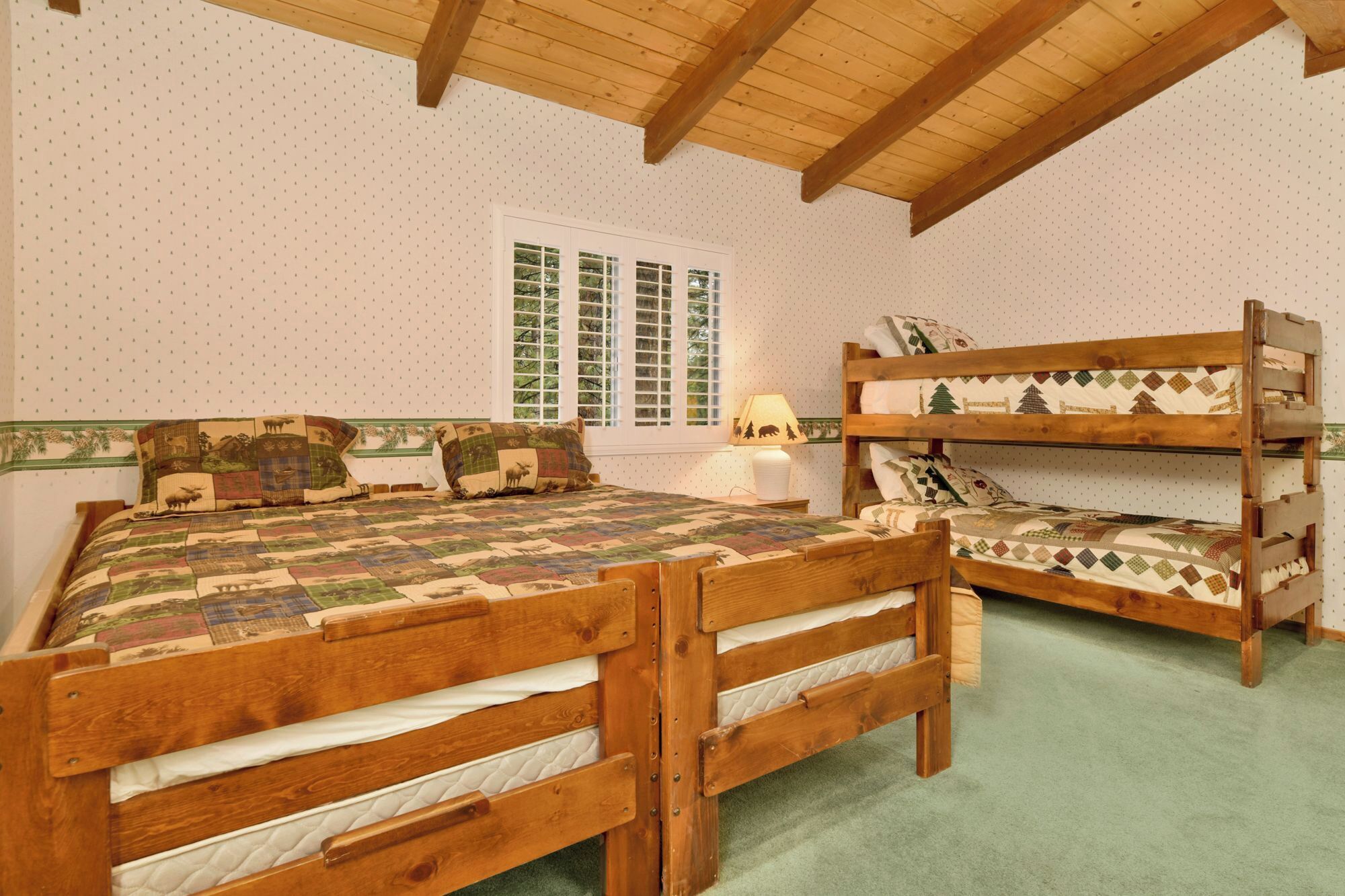 A bedroom with two wooden bunk beds, a quilted bed, a window with blinds, and a lamp on a bedside table.