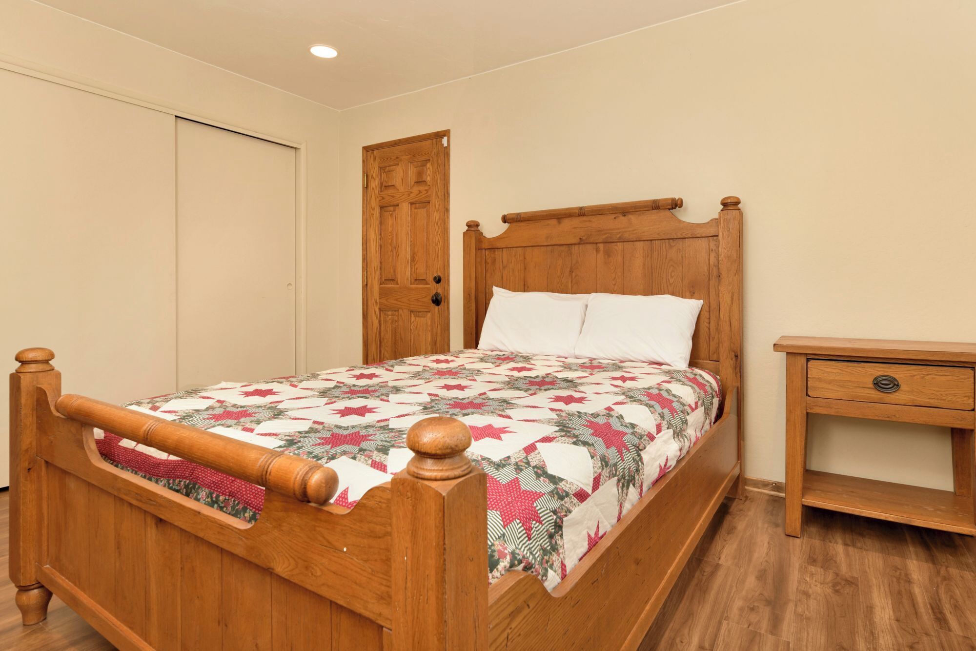 A wooden bed with a quilt, a wooden nightstand, and a door in a room with wooden flooring.