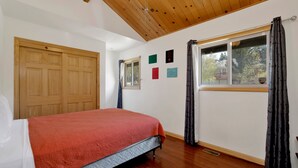 A bedroom with a bed, wooden ceiling, a window with curtains, and a door. - Shasta Mountain Cabin#1916 (Big Bear City)