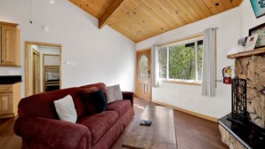 A living room with a wooden ceiling, a red sofa, a wooden table, a stone fireplace, and a window with a view of trees. - Shasta Mountain Cabin#1916 (Big Bear City)