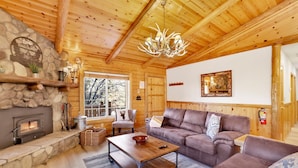 A cozy log cabin interior with a stone fireplace, leather sofas, a wooden coffee table, and a chandelier. - Crescenta Pines LOG Cabin #2346 (Big Bear City)