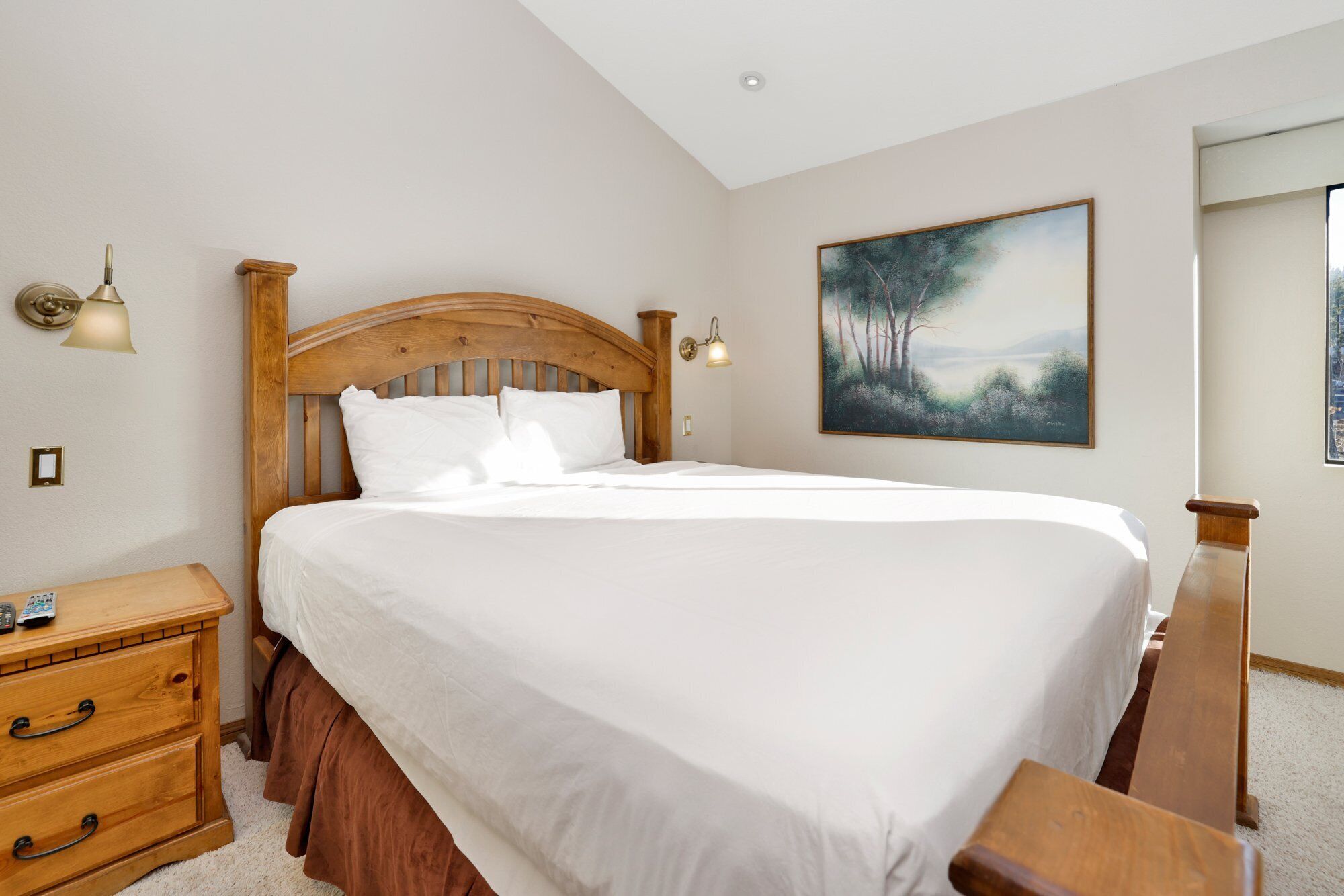Una cama bien hecha en una habitación con cabecera de madera, una mesita de noche con control remoto y un cuadro en la pared.