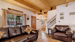 A spacious living room with a brown leather sofa, a smaller leather armchair, and a wooden coffee table. A wooden staircase leads to another room, and a window with curtains is visible. - Whiskey Mountain Lodge - 2028 (Sugarloaf)