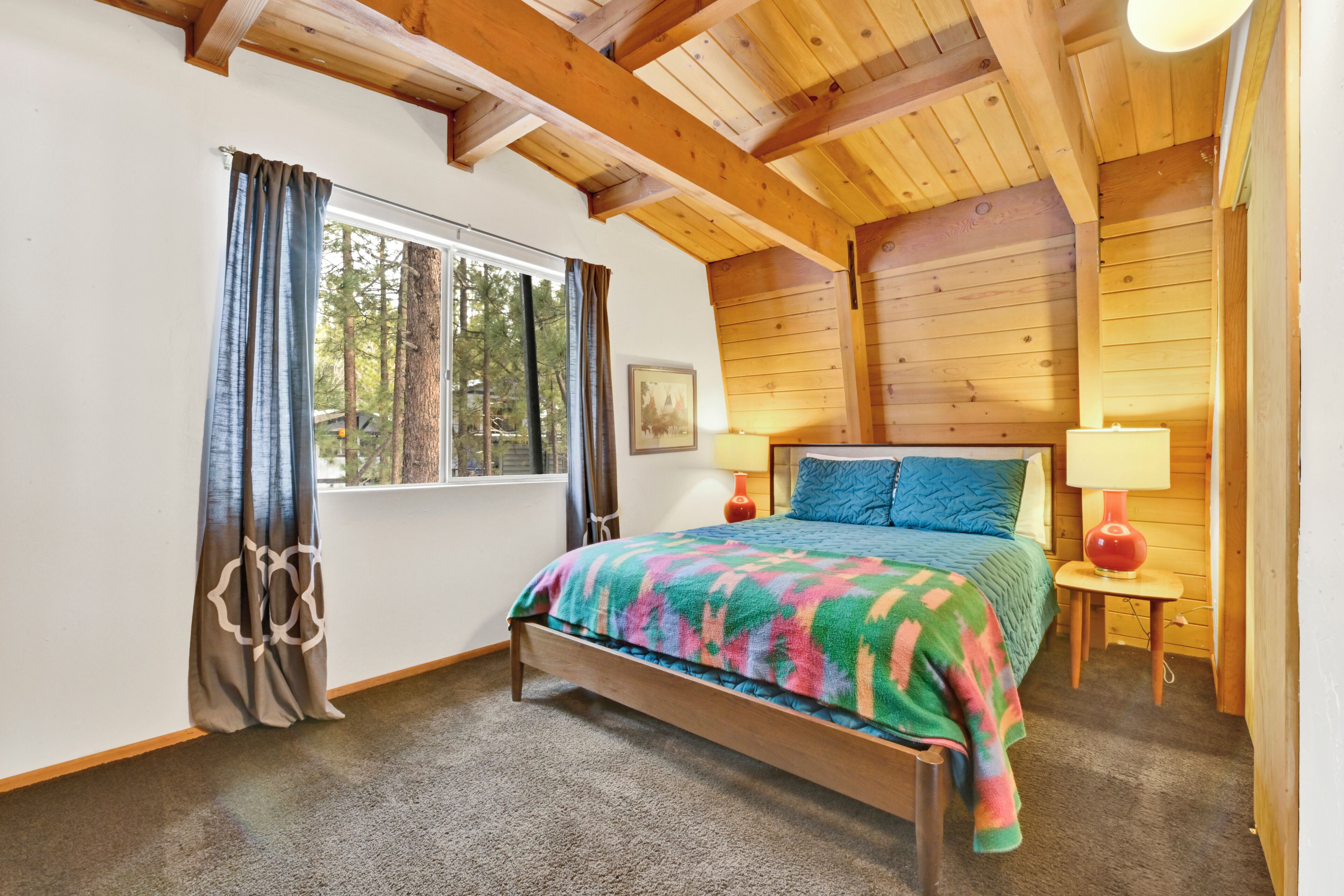 Een slaapkamer met een houten plafond, een groot bed met een kleurrijke deken, twee bedlampjes en een raam met gordijnen.
