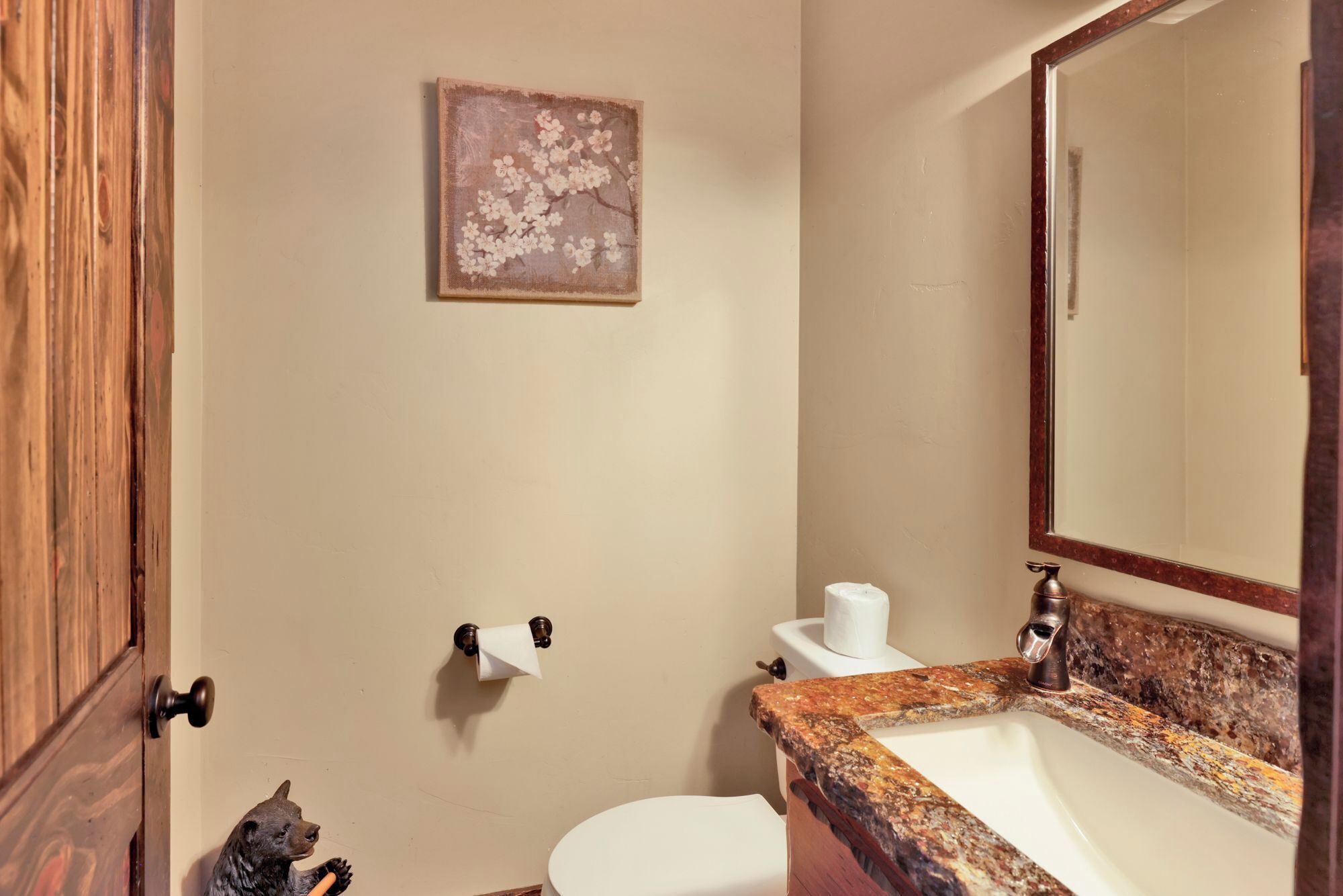 Une salle de bain avec une porte en bois, des toilettes, un lavabo avec un plan de toilette en marbre, un miroir et une œuvre d’art florale encadrée accrochée au mur.