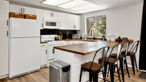 A modern kitchen with white cabinets, a central island with bar stools, a microwave, a stove, and a trash bin.