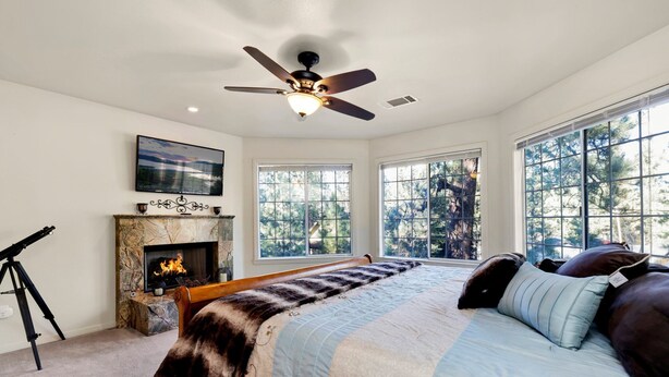 Une chambre avec une cheminée, de grandes fenêtres, un ventilateur de plafond et un lit agrémenté de plusieurs coussins.