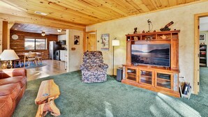 Interior acogedor de una cabaña de troncos con techo de madera, un televisor grande, un sillón estampado y un comedor con mesa y sillas.
